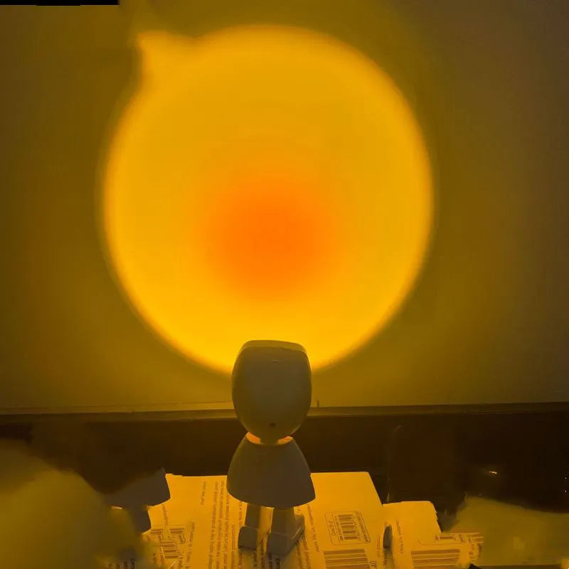 A sunset night light atmosphere lamp with a projection of a golden sunset on a wall, featuring an astronaut figure and illuminating a shelf with flowers and a microphone.