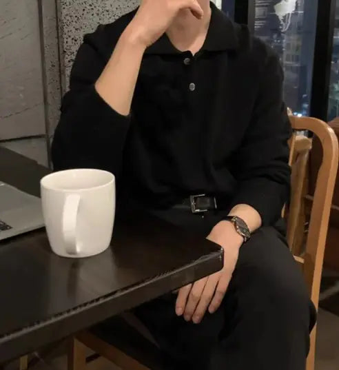 A man sitting at a table wearing a black long-sleeved polo collar buttoned sweater, with a white cup on the table.