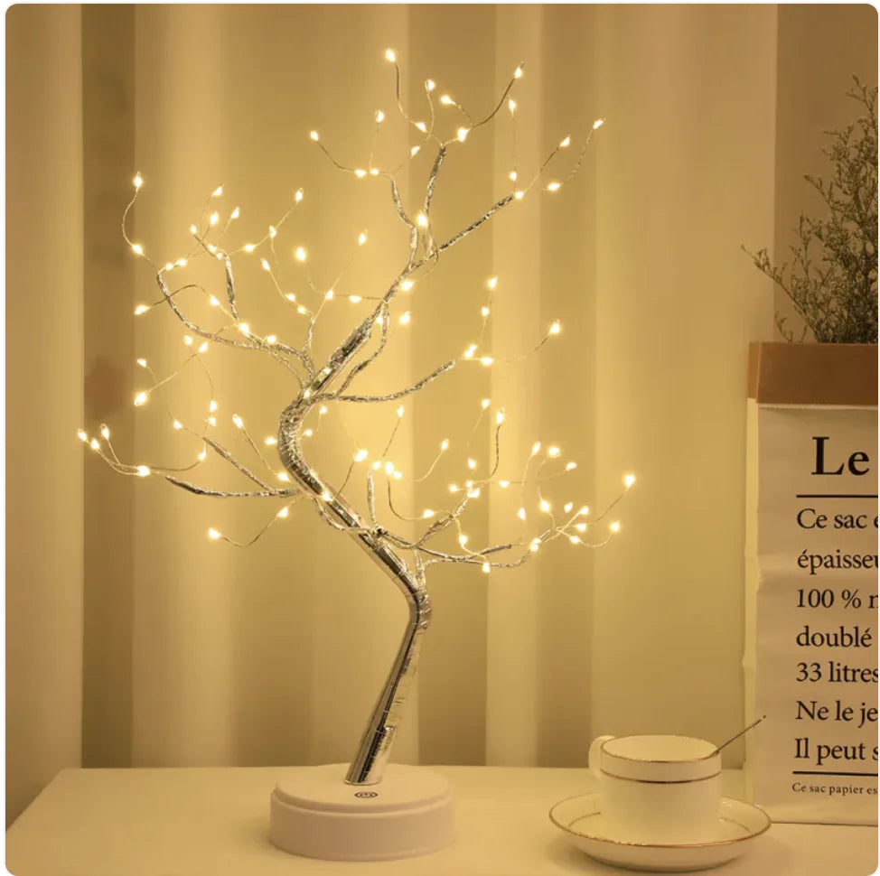 An LED copper wire tree lamp with a starry sky light pattern, placed on a white base, with a neutral warm light color, placed in a room setting with a book and a cup.